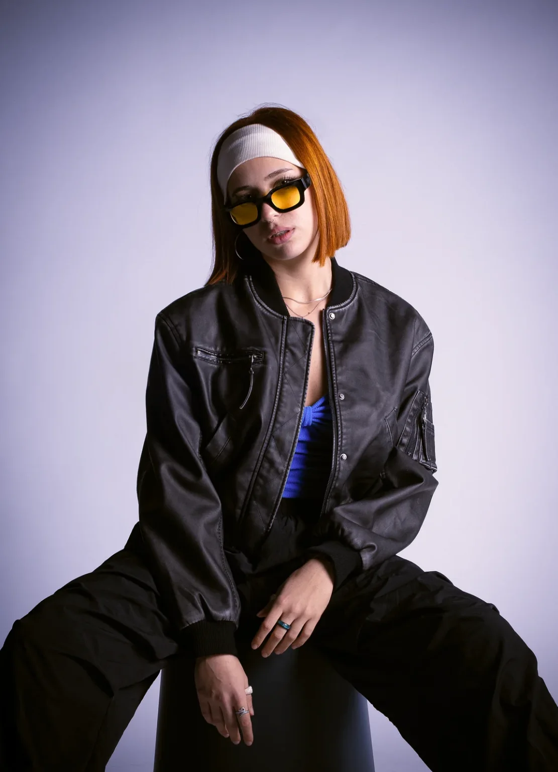 Artista con anteojos amarillos y campera de cuero sentada sobre fondo lila